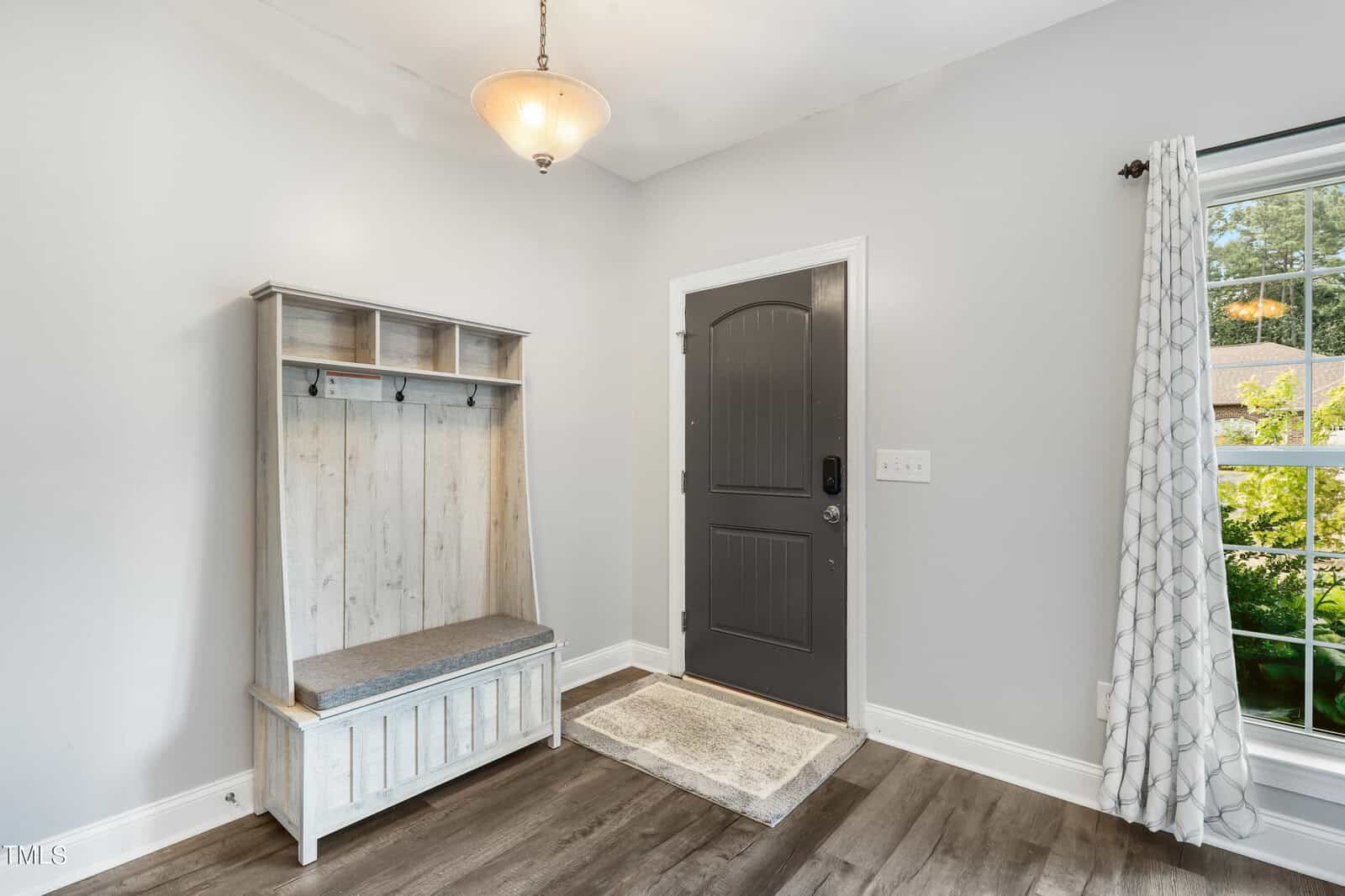 A bright entryway with light gray walls and dark wood flooring. A black door is centered, flanked by a window with patterned curtains. A wooden bench with storage, hooks, and a cushioned seat is on the left. A ceiling light fixture hangs above.