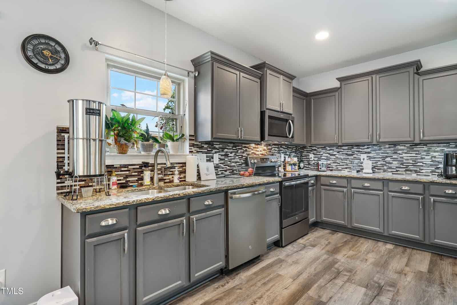 Modern kitchen with gray cabinets, stainless steel appliances, and a granite countertop. A small window with plants above the sink lets in natural light. The backsplash is a mix of gray and white tiles, and the floor features wood-style planks.