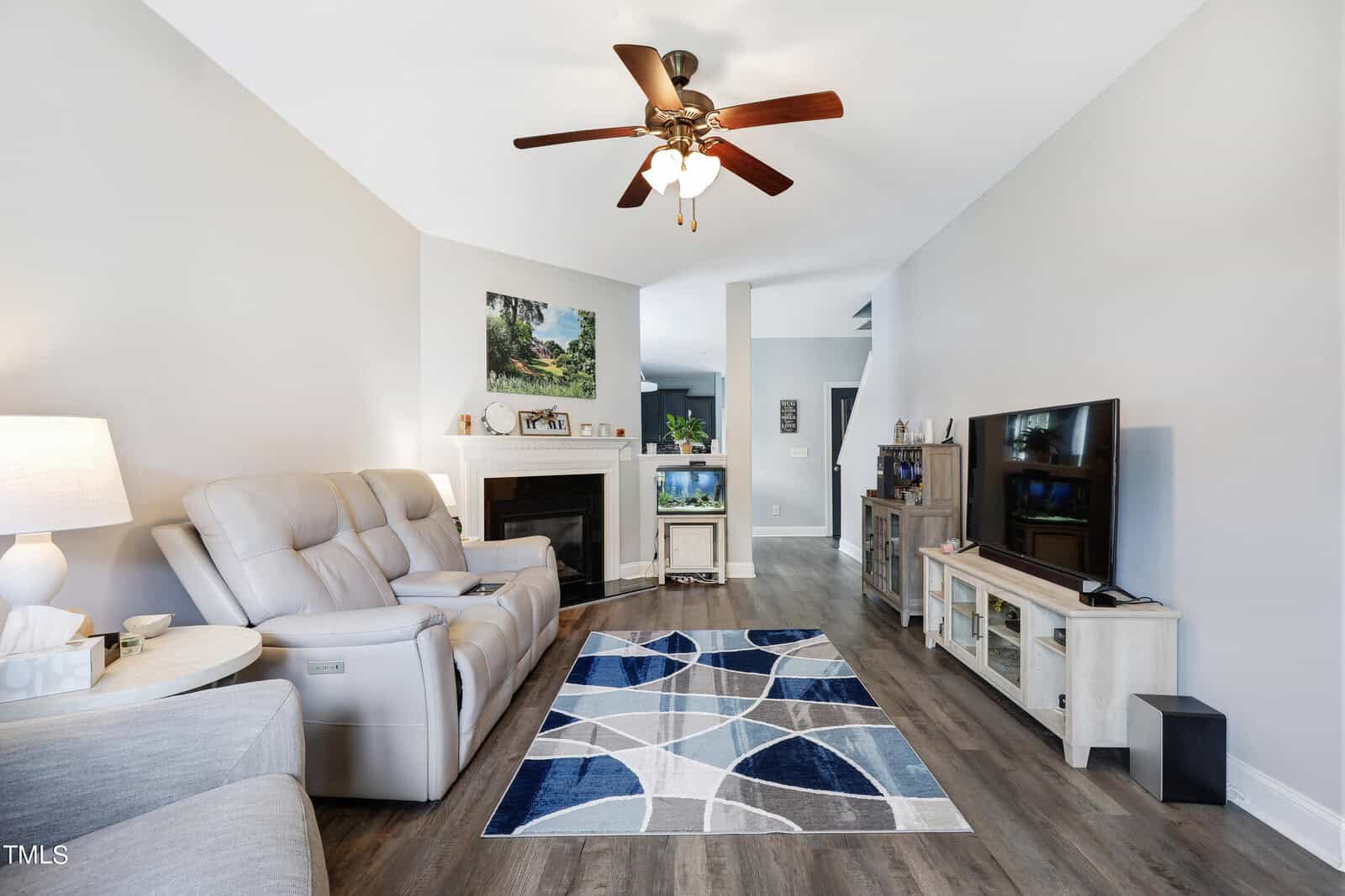 A modern living room with light gray walls and wooden flooring. It features a reclining sofa, a patterned blue and gray rug, a ceiling fan, a TV on a stand, and a fireplace with framed art above. Decorative items are on the shelves and mantel.