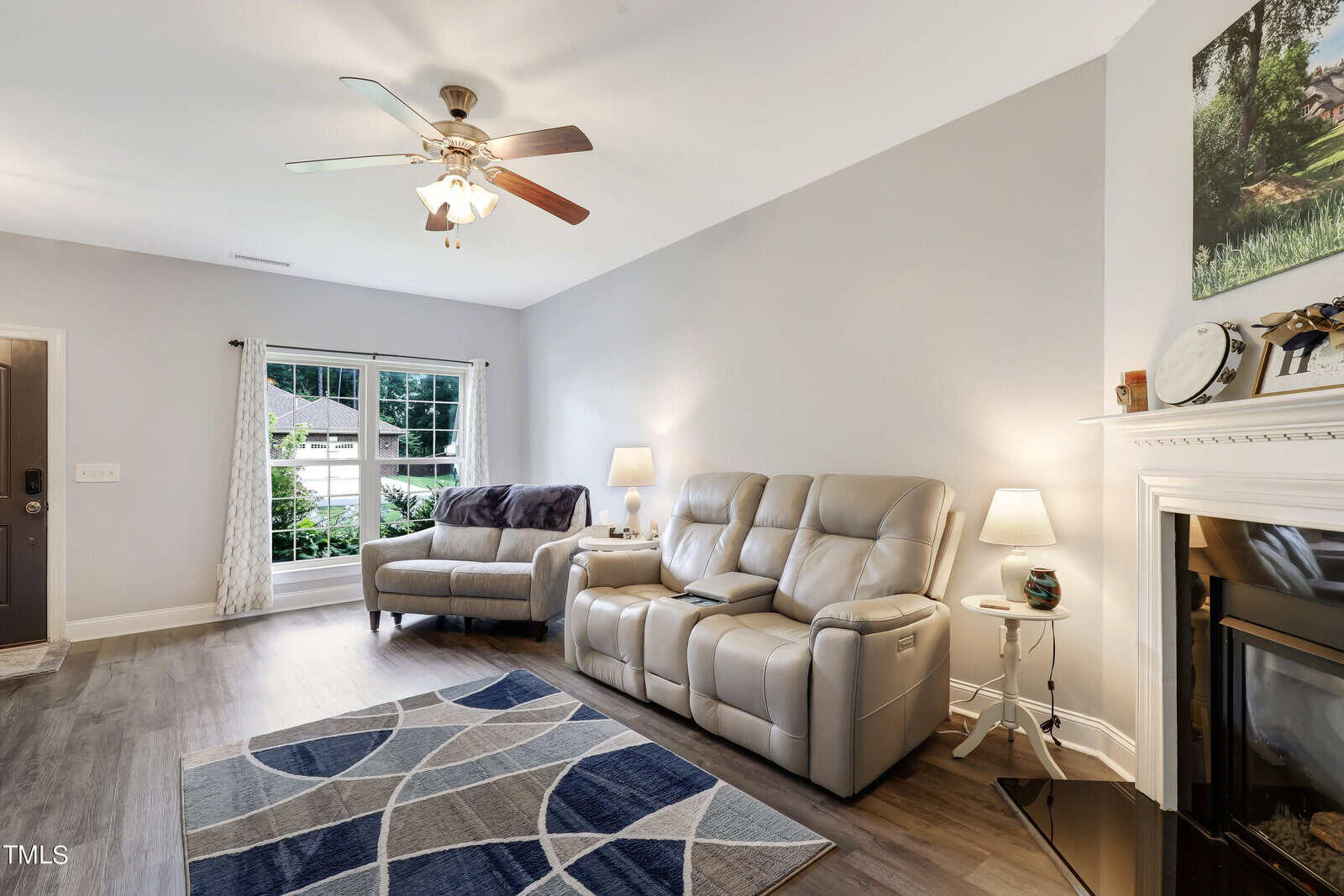 A modern living room with a gray reclining sofa and matching loveseat, a geometric patterned rug, and a ceiling fan. A fireplace is on the right, decorated with a clock and lamp. Large window with sheer curtains offers natural light and a view outside.