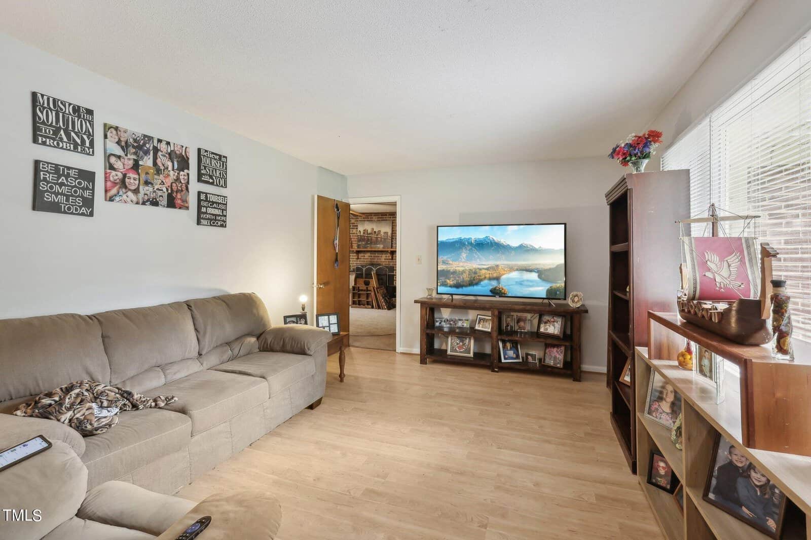 A cozy living room featuring a beige sectional sofa, a flat-screen TV displaying a mountain landscape, framed family photos on the wall and shelves, and a wooden floor. Bright daylight streams in through a window with blinds.
