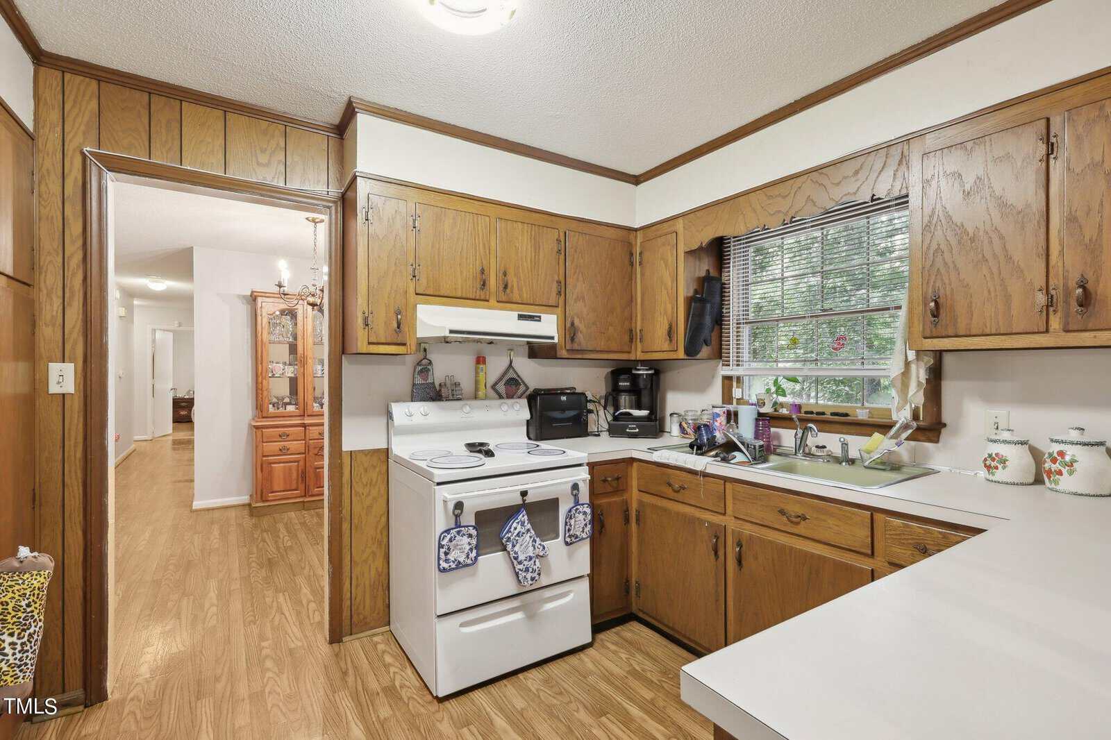 A vintage kitchen featuring wooden cabinets and white countertops. An electric stove with two towels, a coffee maker, and utensils sit on the counter. A window with blinds is above the sink. The floor is wooden, leading to a hallway and another room.