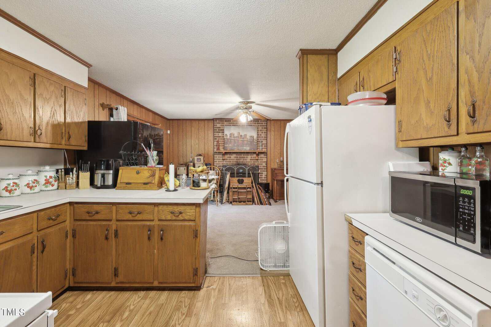 A kitchen with wooden cabinets and white appliances, including a refrigerator, microwave, and dishwasher. A black stove is present. A brick fireplace and fan are visible in the adjoining dining area with a table and chairs.