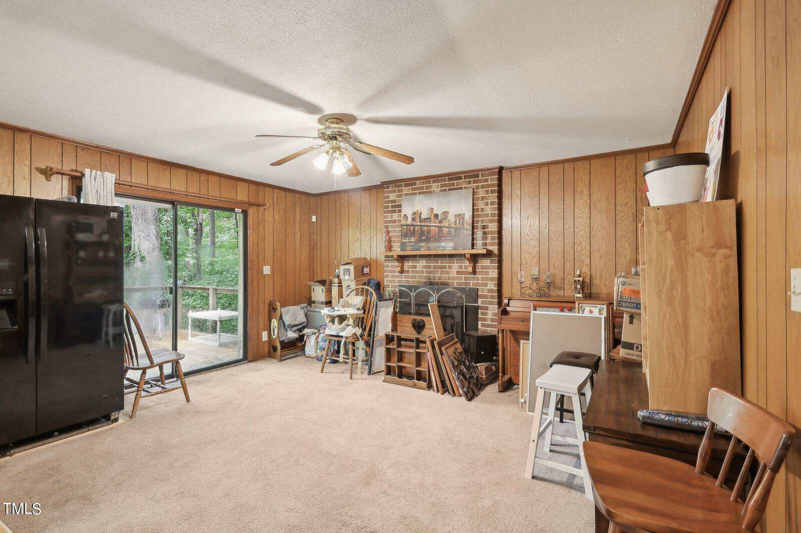 Room with wood-paneled walls, a black refrigerator, ceiling fan, and carpeted floor. Features sliding glass doors to a patio, a brick fireplace, piano, and various wooden chairs and furniture. Cozy and functional space with natural light.