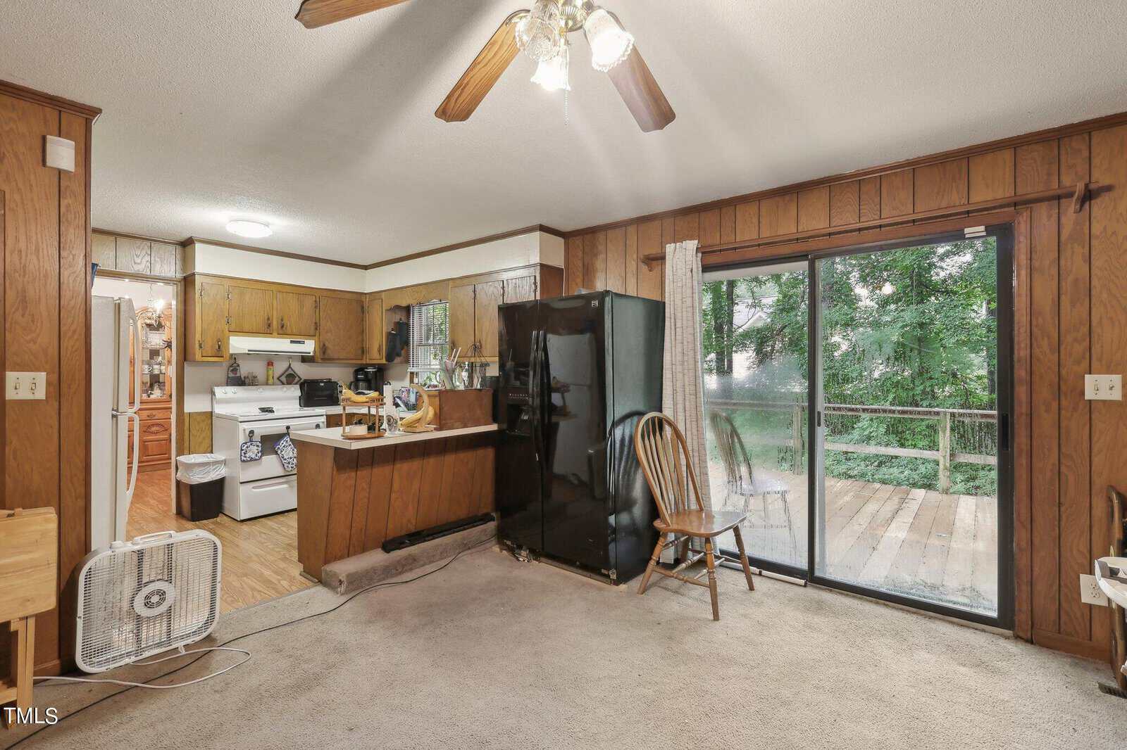 A cozy kitchen and dining area with wooden cabinets, a black refrigerator, and a white stove. A small fan is on the floor. Sliding glass doors lead to a wooden deck outside. A ceiling fan with lights is above.
