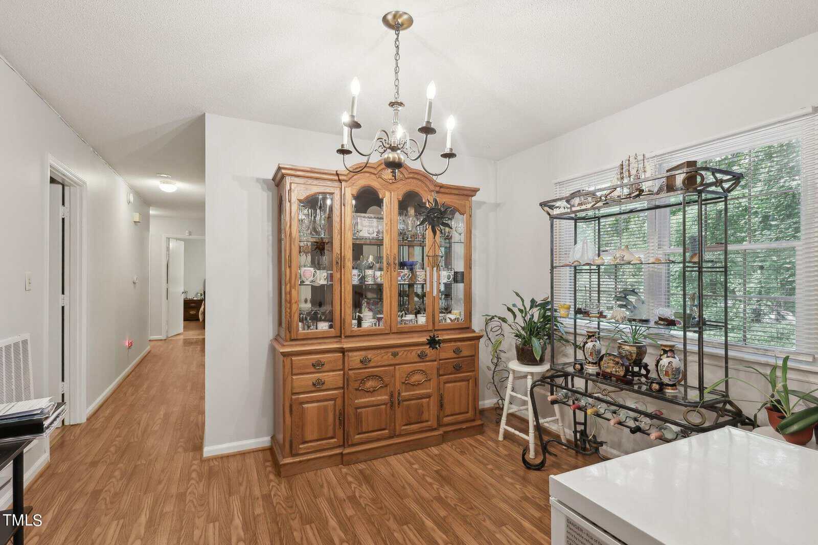 A dining area with a wooden china cabinet displaying glassware and decor. A metal shelf holds plants and knick-knacks by a window. The room features laminate flooring and a chandelier. Hallway visible in the background.