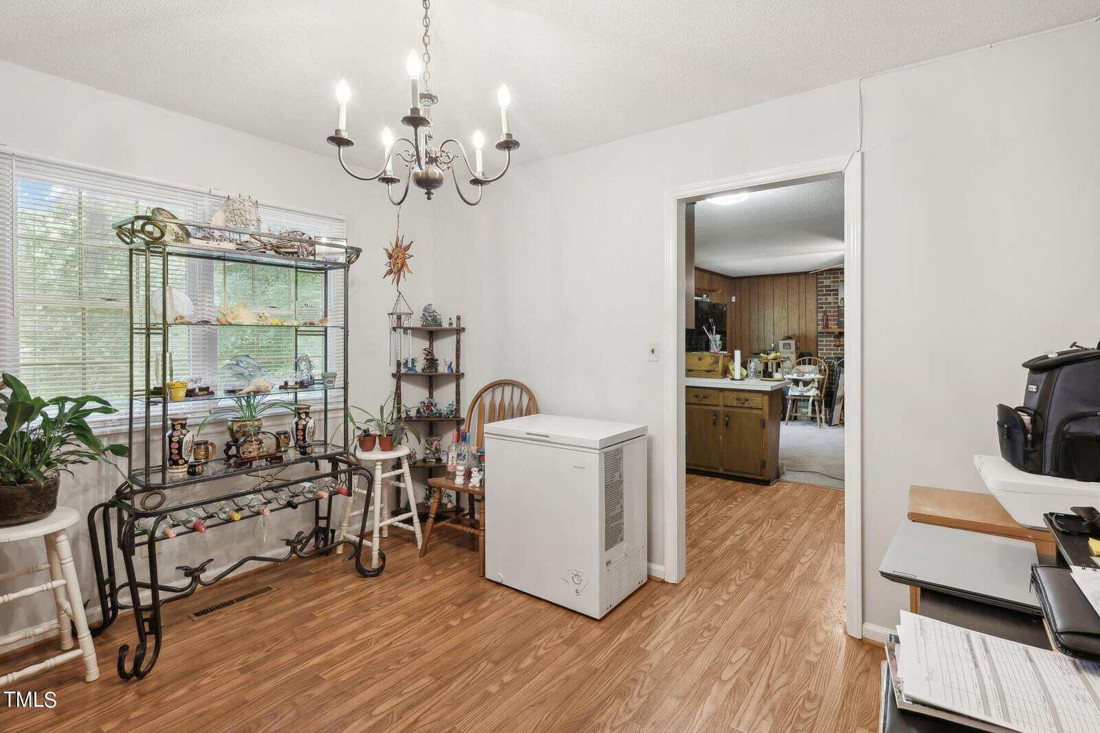 A room with wooden flooring features a chandelier, a window with blinds, and a black metal shelf displaying various items. A white chest freezer stands against the wall, and there's an open doorway leading to another room with a brick wall.