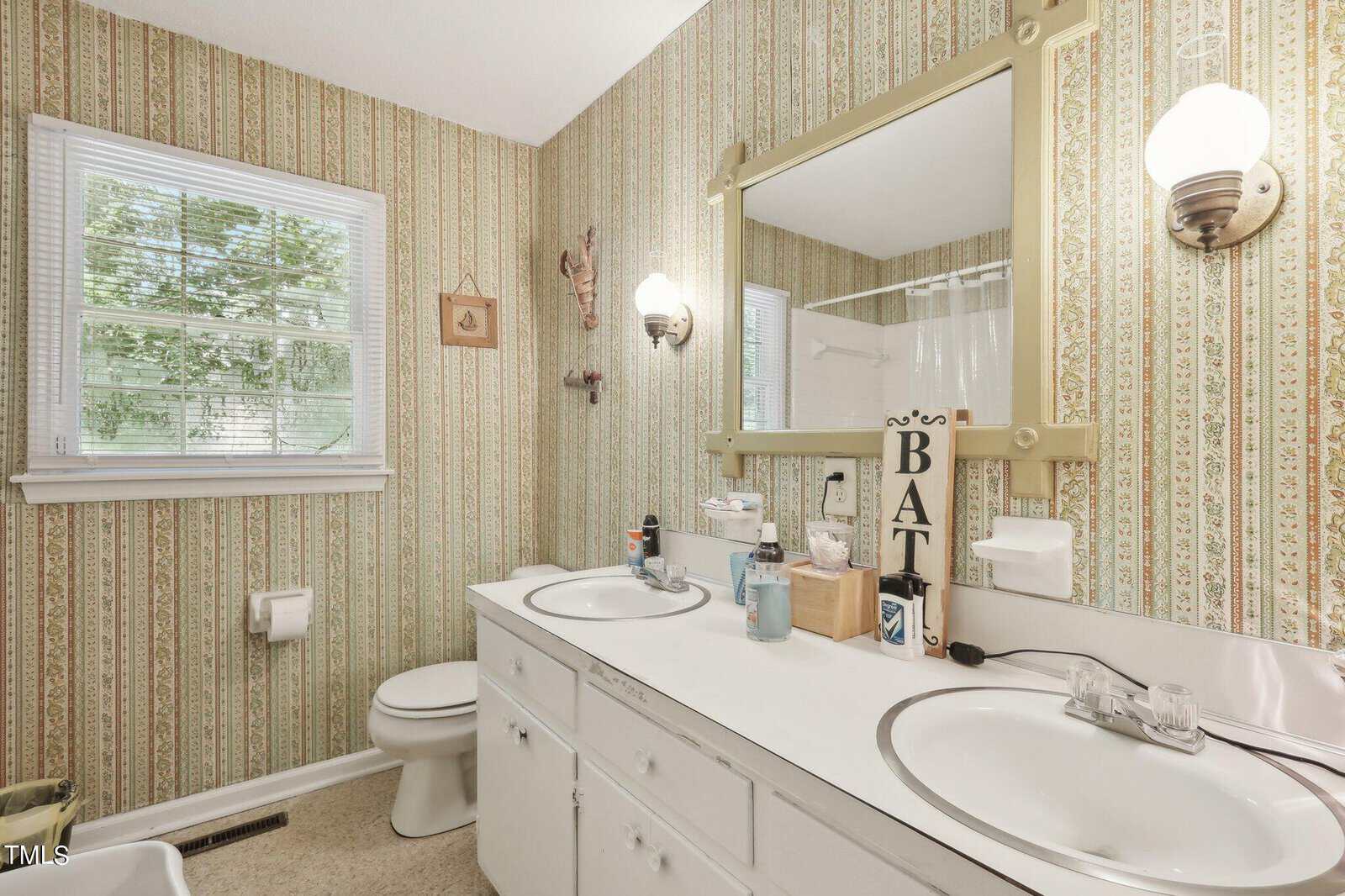 A retro-style bathroom featuring a double sink with a large mirror above. The wallpaper has vertical green and beige stripes. There are two wall-mounted light fixtures, and a window with a view of trees. A "BATH" sign decorates the counter.