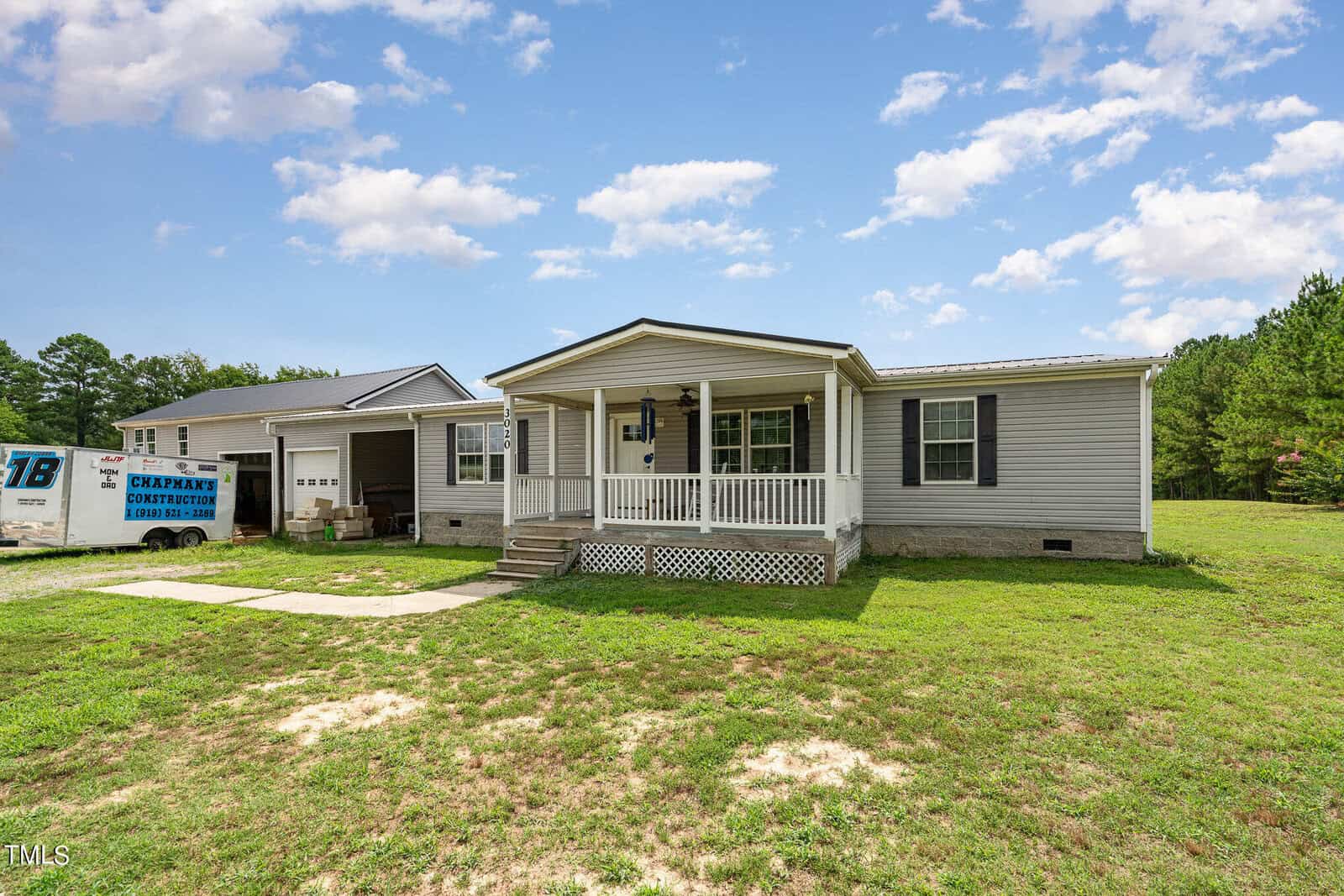A one-story house with gray siding and a front porch sits on a spacious lawn at 3020 Tar River Road, Oxford. A gravel driveway leads to a garage with two vehicles partially visible. A cloudy blue sky is overhead, with trees dotting the background.