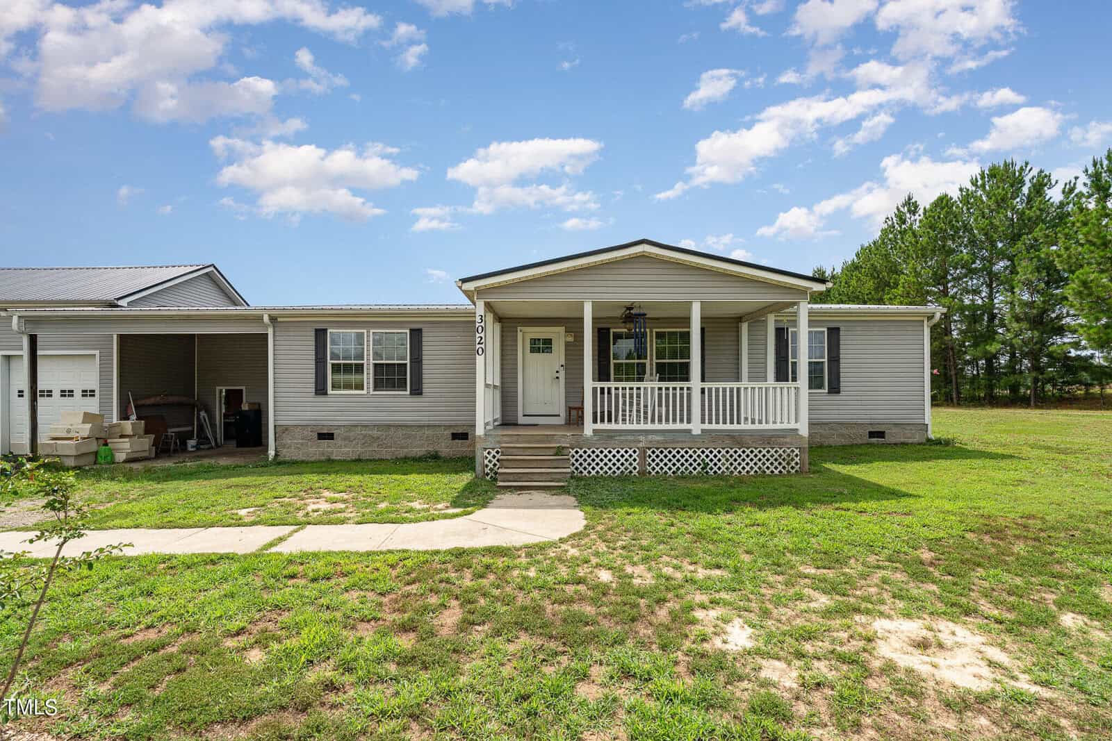 A single-story gray house with a white front porch, steps, and railing. A small driveway leads to an attached garage on the left. The house is set against a grassy yard with trees and a partly cloudy sky in the background.