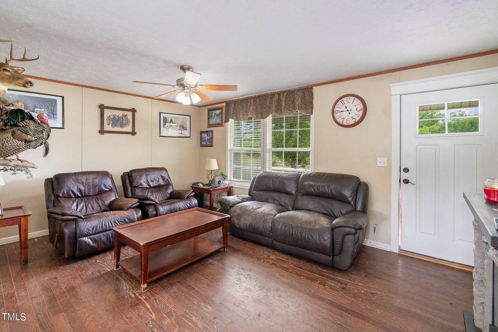 A cozy living room with two recliners, a sofa, and a coffee table on wooden flooring. Walls are adorned with framed pictures and a mounted deer head. A ceiling fan with lights hangs above, and a window lets in natural light. A clock is near the door.