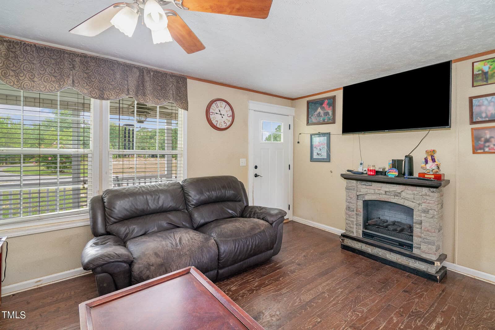 A cozy living room featuring a leather sofa, a wall-mounted flat-screen TV above a stone fireplace, and several framed pictures. The room has a wooden coffee table, a ceiling fan, and large windows with curtains, letting in natural light.