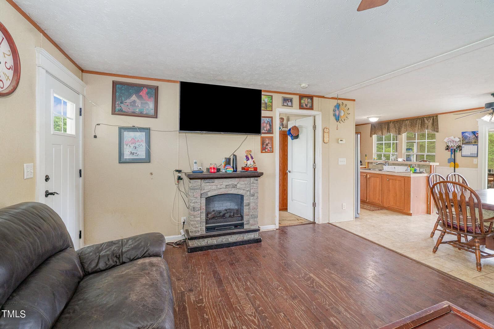 A cozy living room with a dark sofa and a wall-mounted TV above a decorative fireplace. The room features wooden flooring and connects to an open kitchen with wooden cabinets and a dining table. Various art pieces and photos decorate the walls.