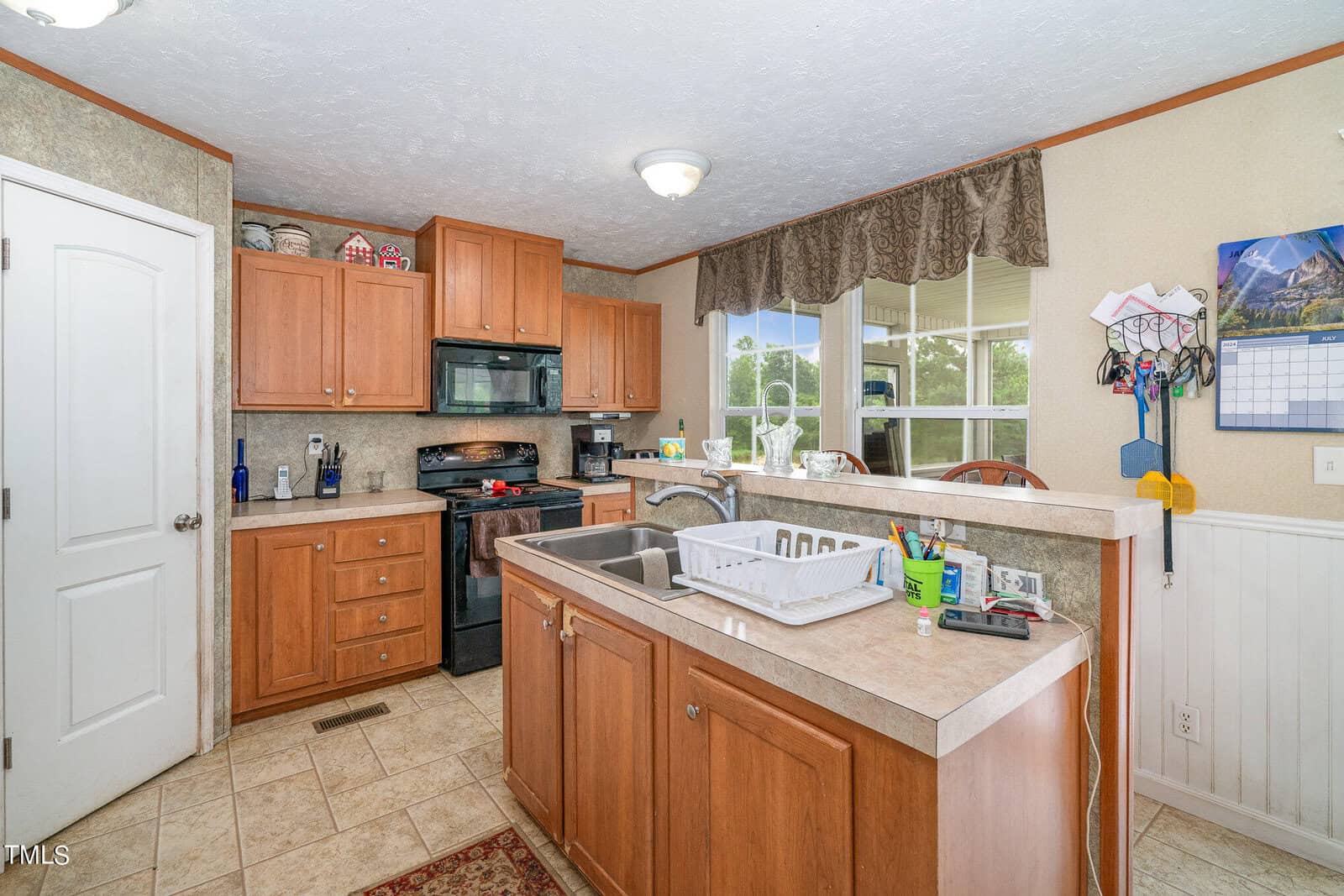 A cozy kitchen with wooden cabinets, a double sink, and black appliances. A drying rack sits by the sink. A window with a view outside adds natural light. A calendar and coat rack are on the wall, and various kitchen items are on the counter.
