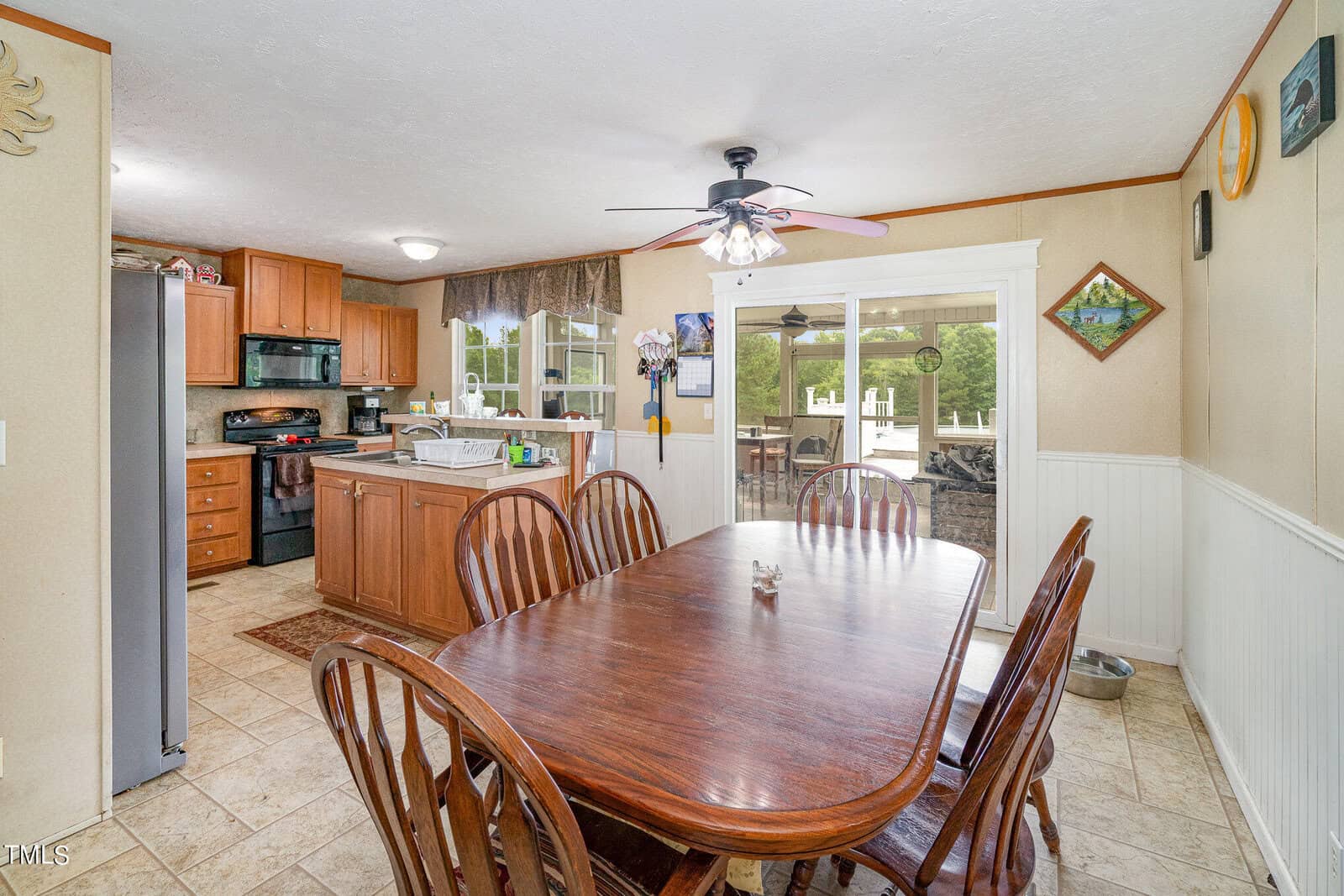 A spacious kitchen with wooden cabinets, black appliances, and a tiled floor. A large wooden dining table with eight chairs is in the foreground. A ceiling fan with lights hangs above, and a sliding glass door opens to a patio outside.