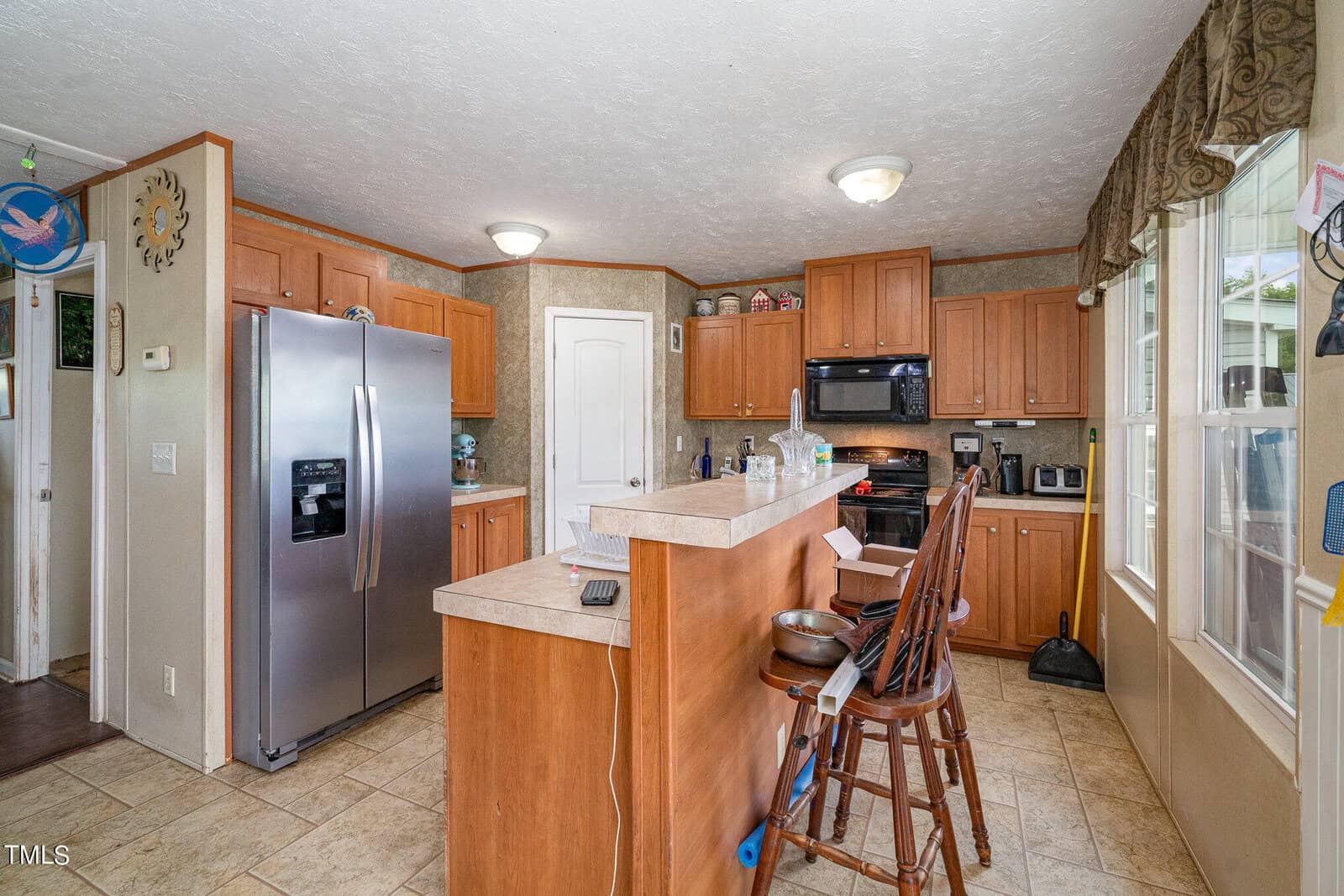 A kitchen with wooden cabinets, a silver refrigerator, and black stove. A small island bar with two wooden chairs is in the center. The floor is tiled, and sunlight enters through a large window on the right.
