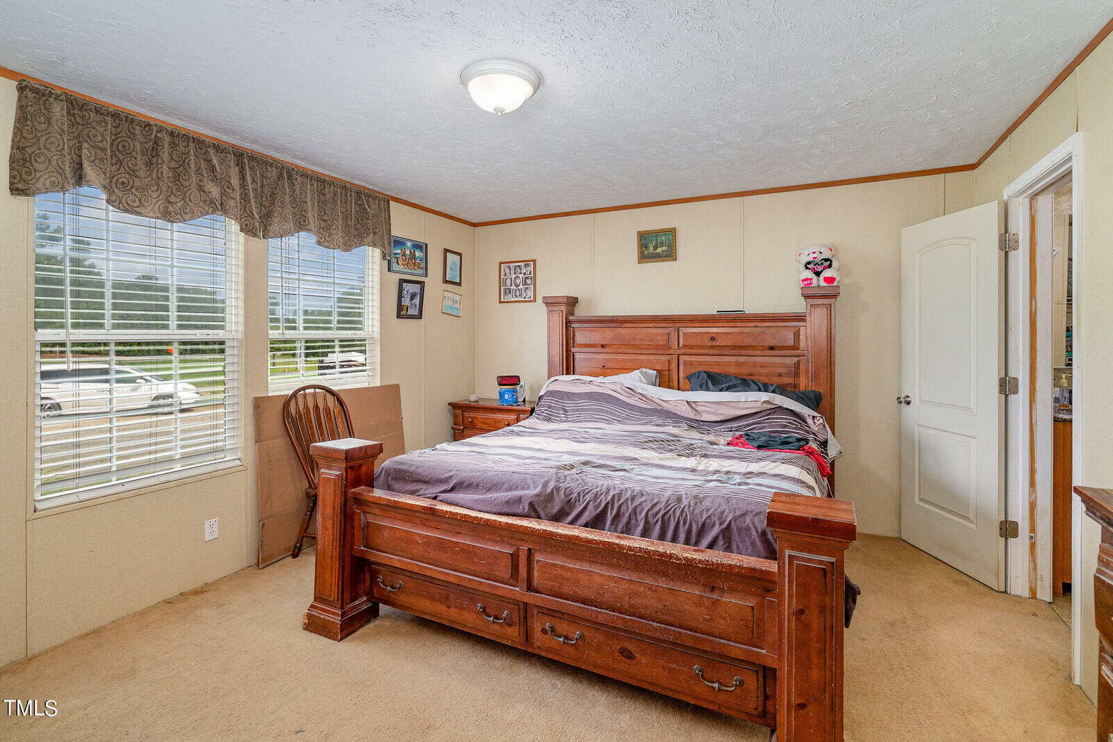 A cozy bedroom with a large wooden bed and nightstand. The bed is unmade, and a stuffed animal sits on the corner. Several framed pictures hang on the walls. Multiple windows let in natural light, and the carpet is a light beige color.