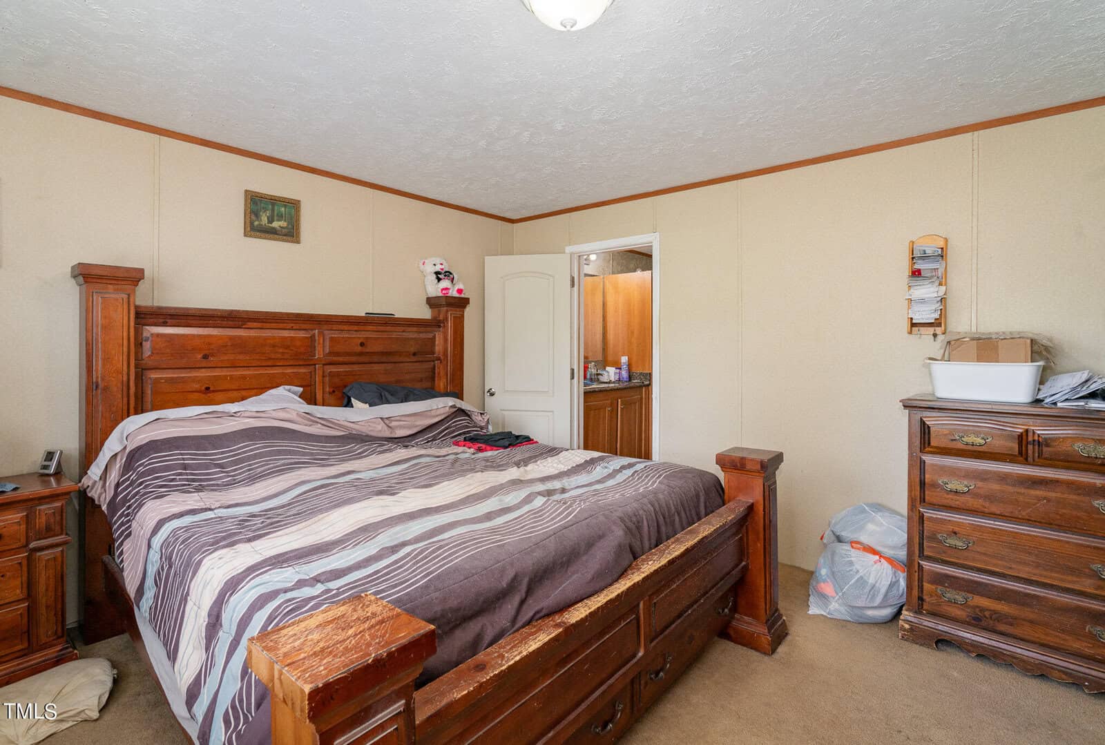 A cozy bedroom features a large wooden bed with striped bedding, flanked by wooden nightstands. A cluttered dresser is on the right, and an open door reveals a bathroom. A small shelf with mail hangs on the wall.