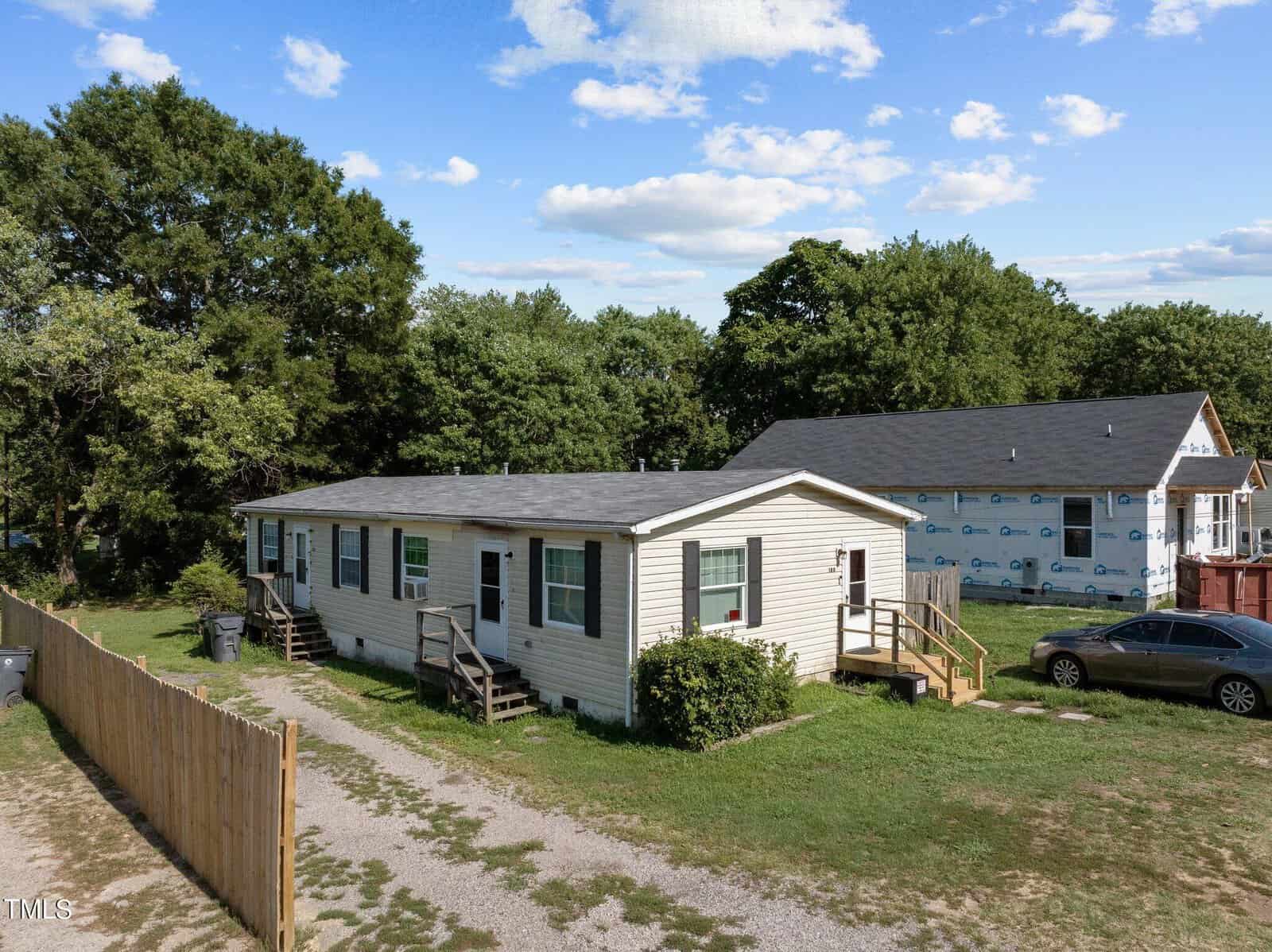 Located at 120 Daniel Street in Oxford, this one-story light-colored house features a dark roof and is surrounded by lush grass and trees. A charming gravel path leads to the entrance, with a car parked nearby. In the background, another house is under construction.