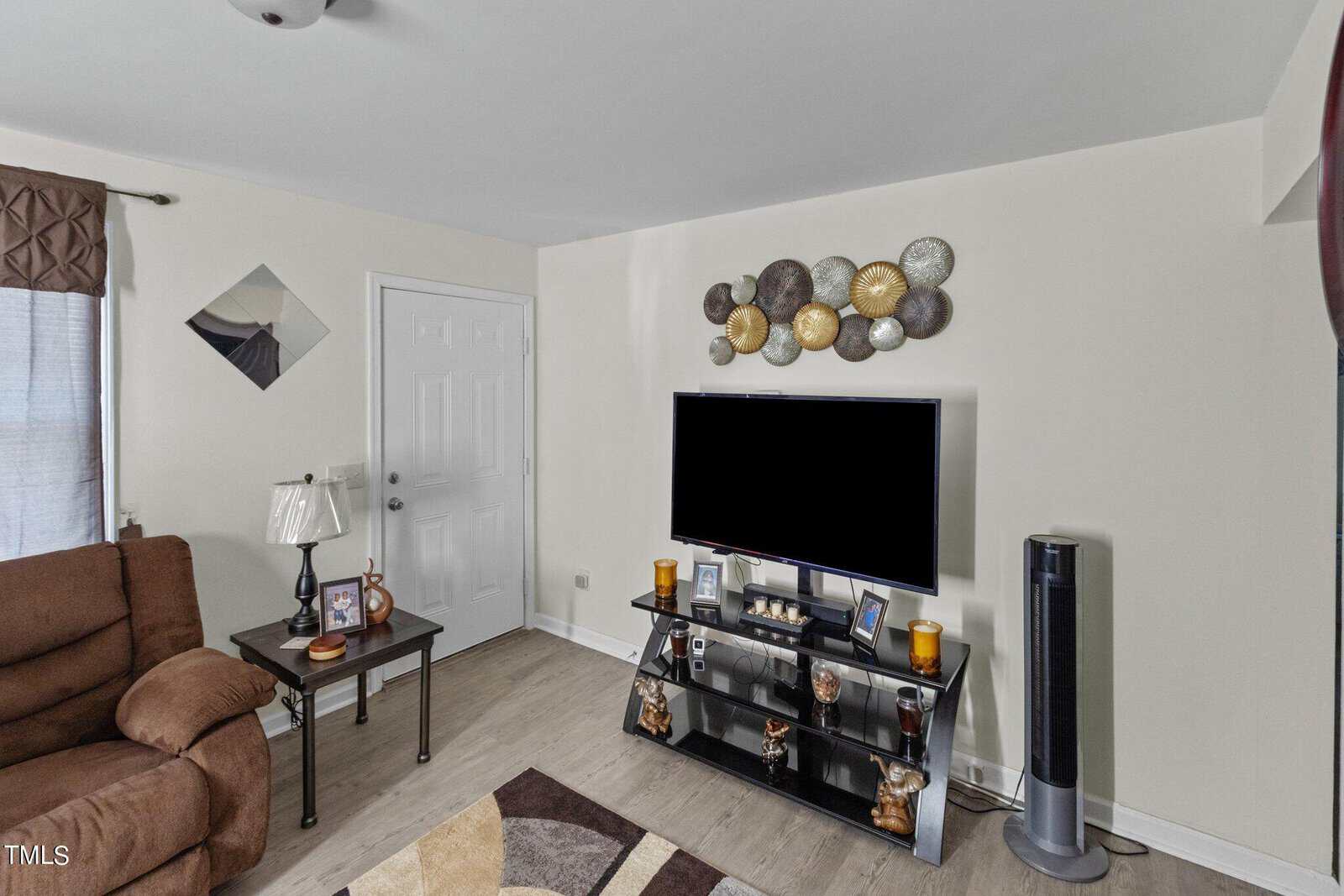 A living room featuring a wall-mounted TV above a media stand with decorative items. A brown recliner, side table with lamp, and wall art are visible. A tall fan stands nearby. The decor includes earthy tones and varying textures.
