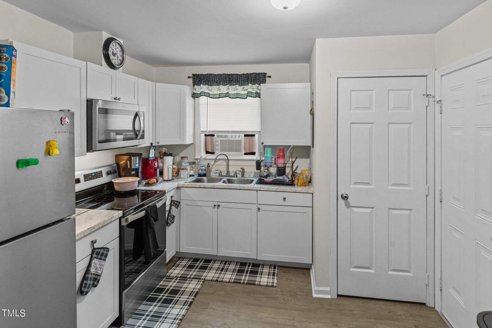 A compact kitchen with white cabinets, an electric stove, a microwave, and a stainless steel refrigerator. A small window with a green curtain sits above the sink. The floor features a checkered rug. Various kitchen items are on the counters.
