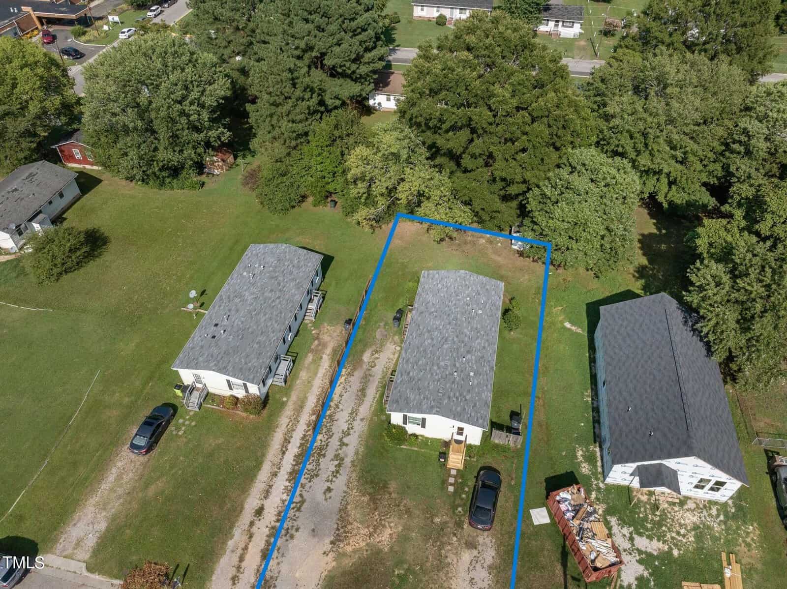 Aerial view showing three small white houses with gray roofs. The house in the center is outlined in blue. There are cars parked on the lawns and trees in the background. .
