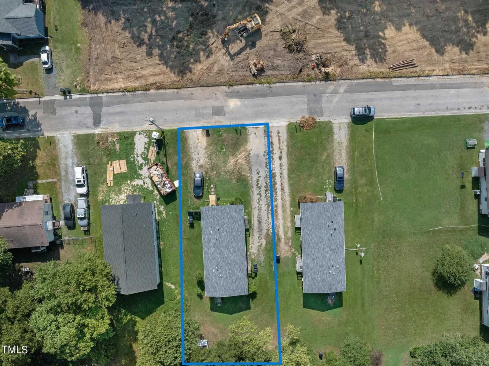 Aerial view of two identical houses inside a blue-outlined rectangular lot, separated by a paved driveway. Each house has a gray roof and is surrounded by grass. Nearby, cars are parked on the street and adjacent properties. Construction equipment is visible at the top.