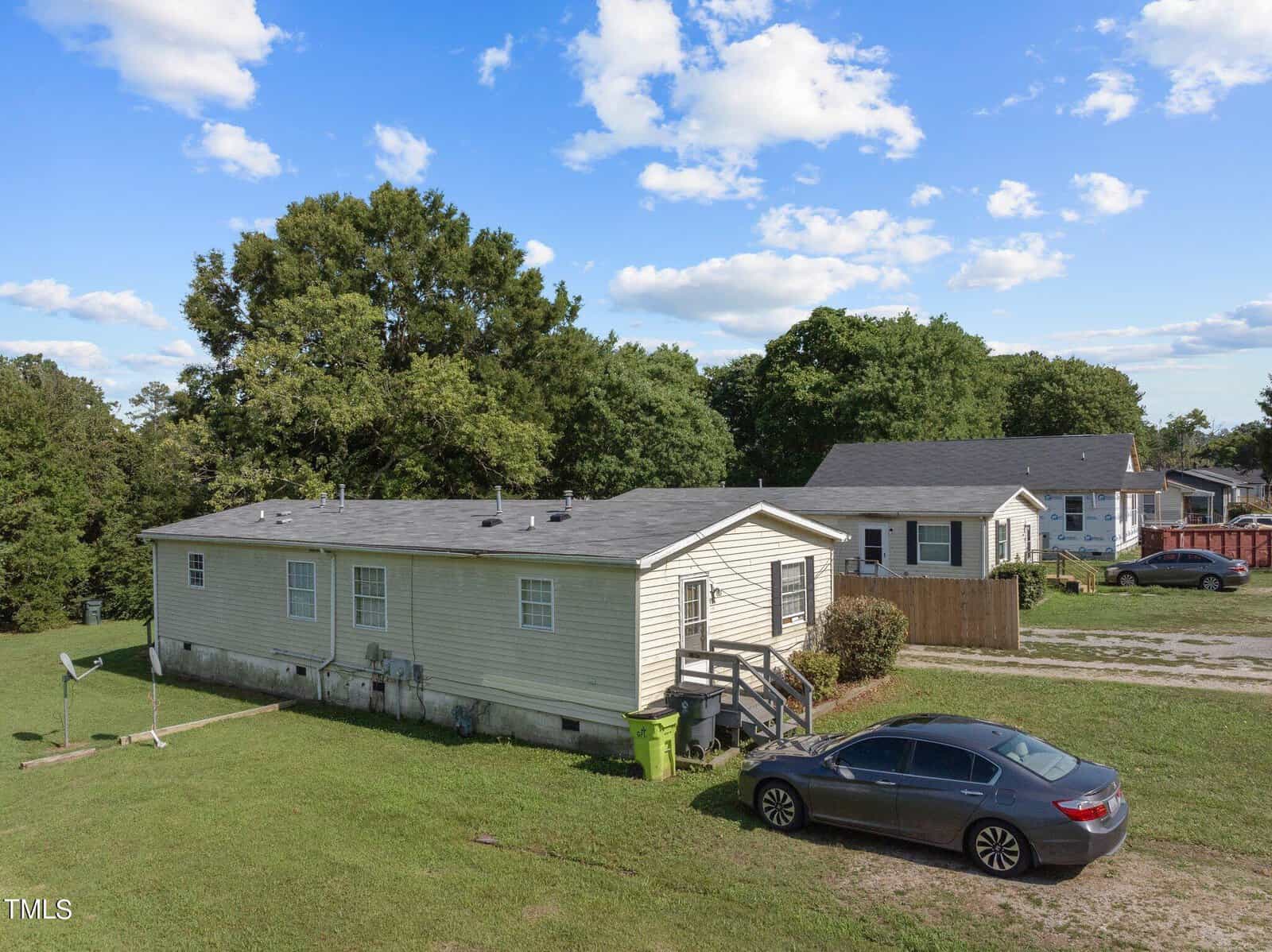 A single-story mobile home with light-colored siding is situated on a grassy lot. A gray car is parked in front of the house. Trees and other houses are visible in the background under a blue sky with scattered clouds.