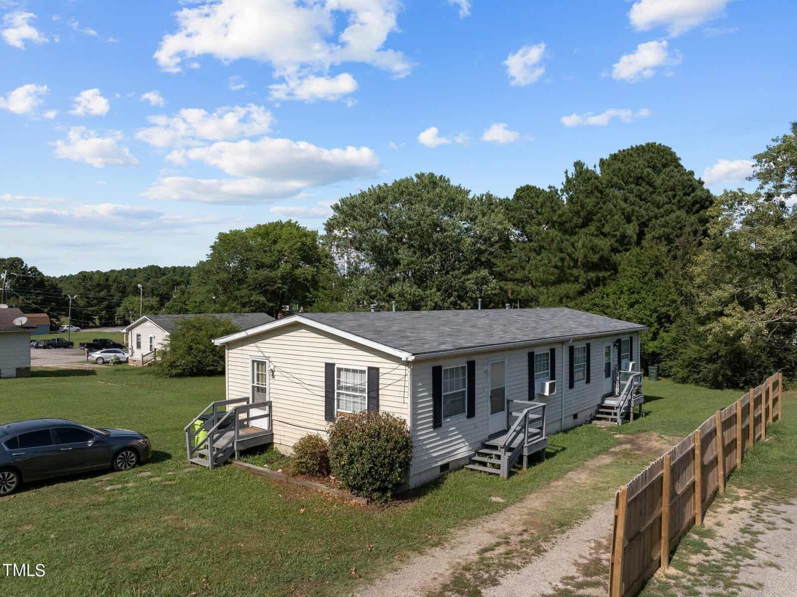 A single-story beige mobile home with two small wooden porches on either side, surrounded by a grassy area and trees. A gray car is parked nearby. The sky is partly cloudy, and there's a wooden fence along the gravel driveway.