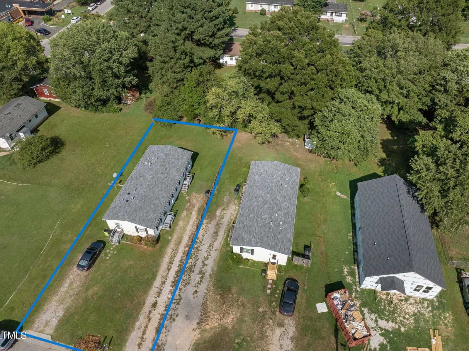 Aerial view of three houses with gray roofs surrounded by trees and grass. One house has a blue outline around it. Two driveways have parked cars. A dirt path runs between the houses and the trees.