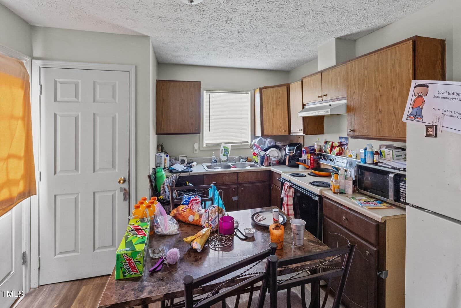 A cluttered kitchen with wooden cabinets, a stove, and a microwave. The counter is crowded with various grocery items, cups, and dishes. A window with blinds is partially open, and a poster is taped to one of the cabinets.