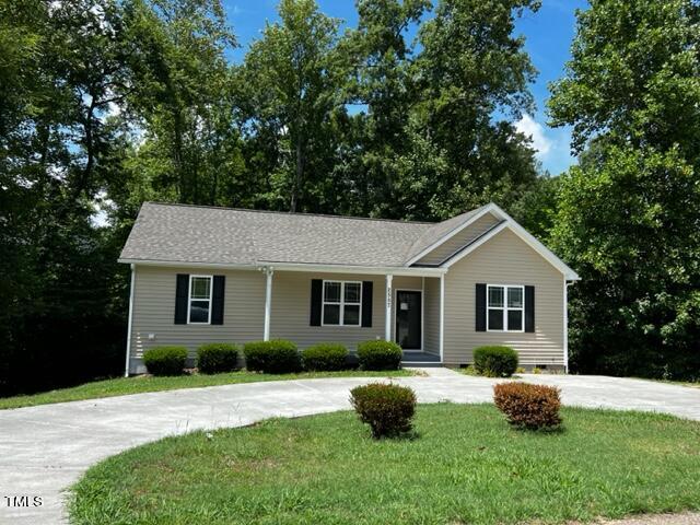 A small, single-story house with beige siding and a gray roof sits in front of a lush, wooded area. The house features a black front door and shutters, with a neatly trimmed lawn and a curved concrete driveway in the foreground.