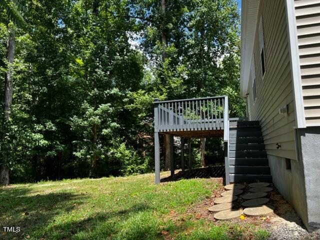 A side view of a house with a raised wooden deck and stairs leading down to a grassy backyard. The area is surrounded by tall, green trees under a clear blue sky.