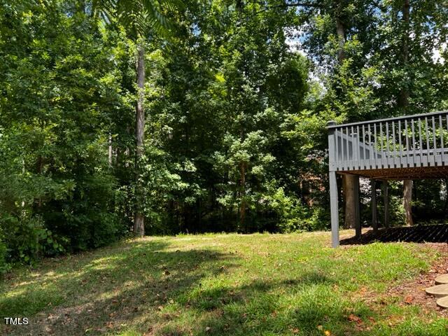 A wooden deck overlooks a grassy backyard bordered by dense trees. Sunlight filters through the foliage, casting dappled shadows on the lawn and creating a serene, natural setting.