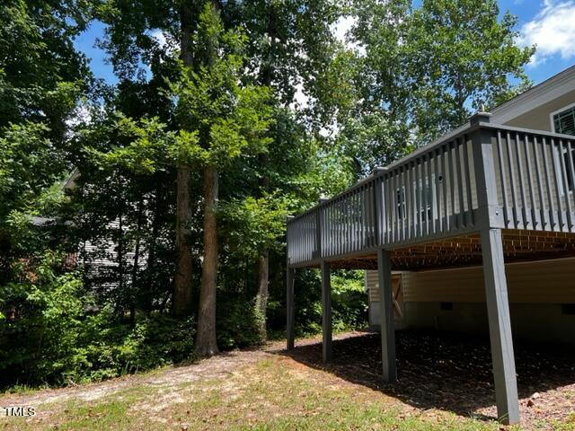 Elevated wooden deck with a gray railing surrounded by tall green trees. The sky is partly cloudy, and the ground below the deck is covered with dry leaves and sparse grass. A house is partially visible behind the deck.