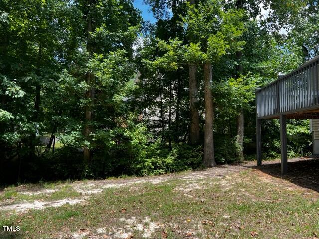 A patch of grass and dirt in a wooded area with tall green trees. A wooden structure, possibly part of a deck, is visible on the right edge of the image. The sky is partly cloudy with bright blue patches.