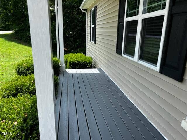 A side view of a house porch with gray wooden flooring and white columns. The house has beige siding and black shutters. Bushes are neatly trimmed along the edge of the porch, with grass visible in the background.