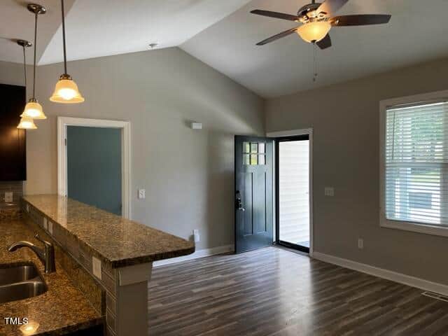 A modern room with a vaulted ceiling, featuring a granite countertop island with a sink, pendant lights, a ceiling fan, and large windows. The open door allows natural light to illuminate the space, highlighting the wood-patterned floor.
