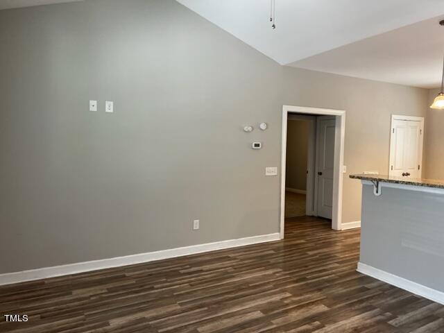 A room with light gray walls and wooden flooring. The ceiling is vaulted. There is a doorway leading to another room and a partial view of a kitchen counter on the right. Light switches and a thermostat are mounted on the wall.
