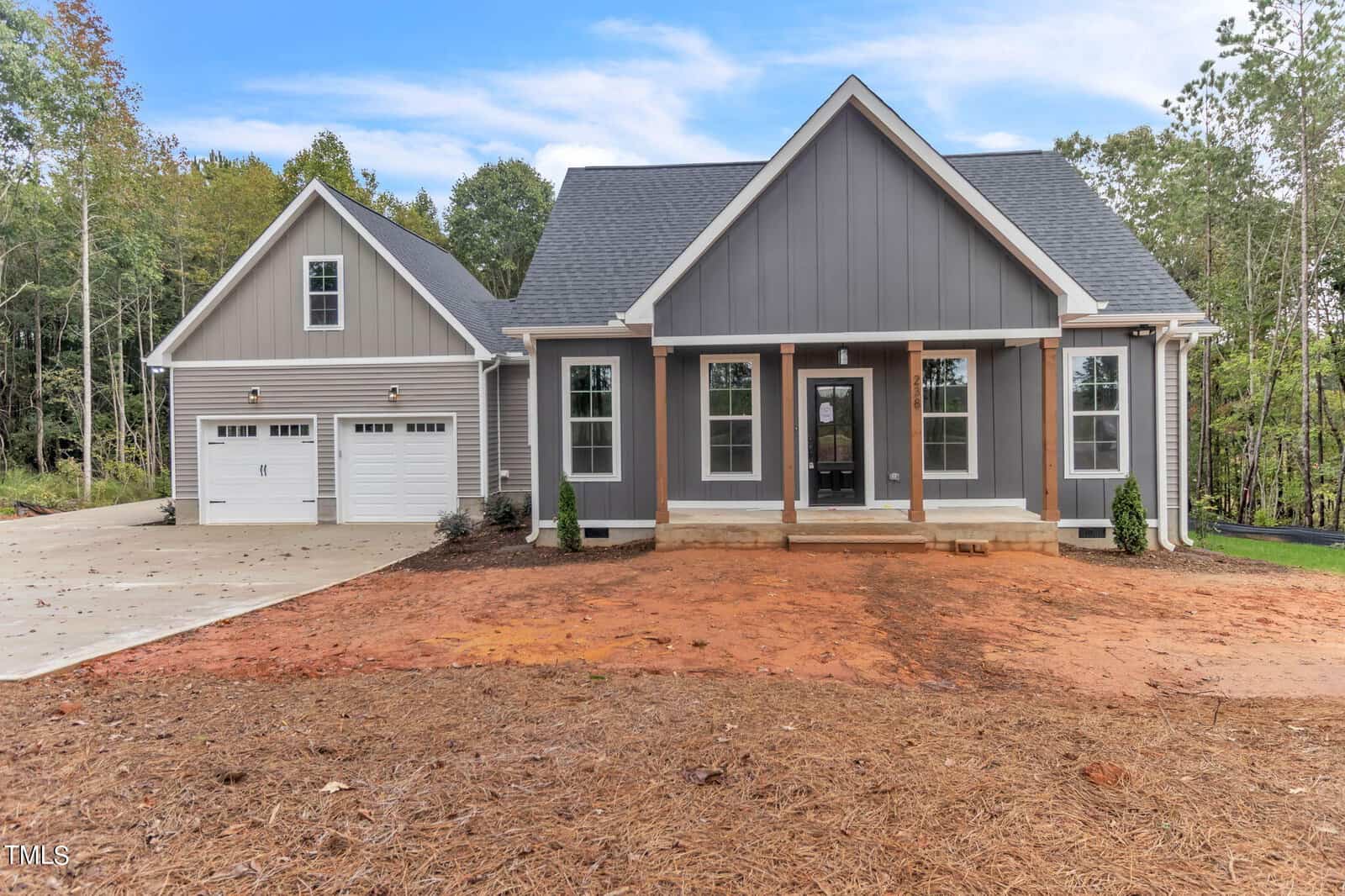 A modern, gray single-family home with white trim and a spacious front porch located at 238 Cheeks Quarter Road in Henderson. The house boasts multiple large windows, a gabled roof, and an attached two-car garage on the left. The front yard features a dirt and mulch area, surrounded by trees and a clear blue sky.