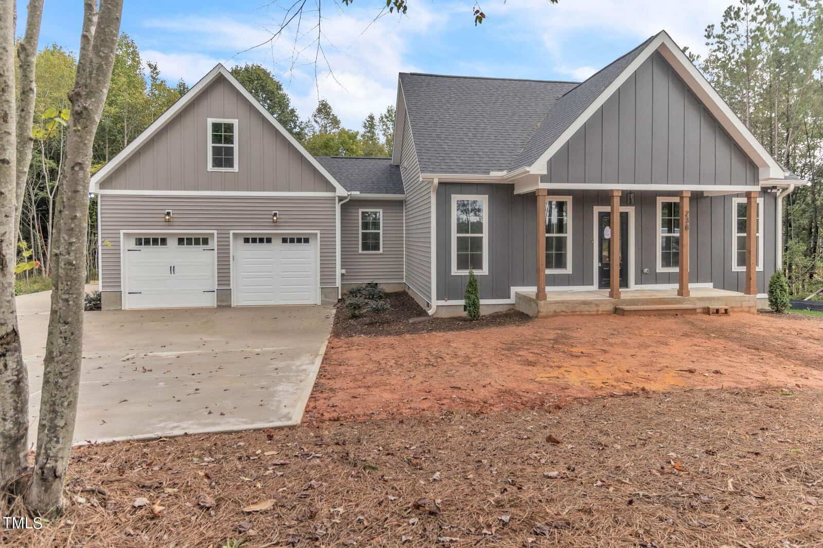 A modern, single-story gray house with white trim featuring a two-car garage, a covered front porch with columns, and large windows. The house is set against a backdrop of trees, with a dirt front yard and a concrete driveway.