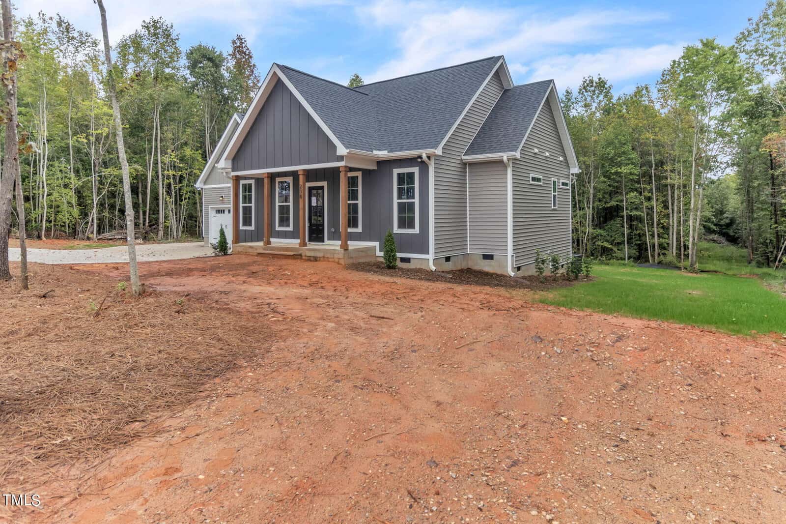 A single-story house with a gable roof and gray siding is surrounded by a partially wooded area. It has a covered front porch and a driveway on the left leading to a garage. The yard is a mix of red earth and patches of green grass. It's a sunny day with a blue sky.
