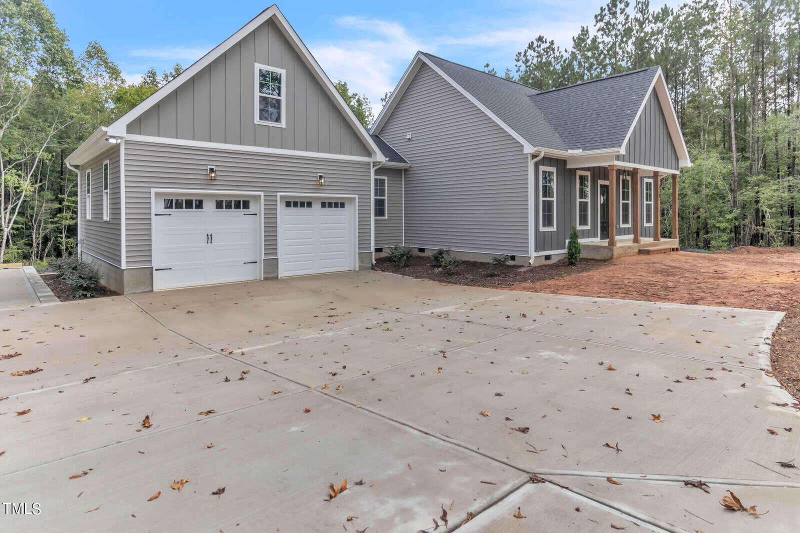 A modern grey two-story house with white trim, featuring two garage doors on the left. The property has a spacious concrete driveway and is surrounded by a wooded area with tall trees. The sky is clear with some clouds.
