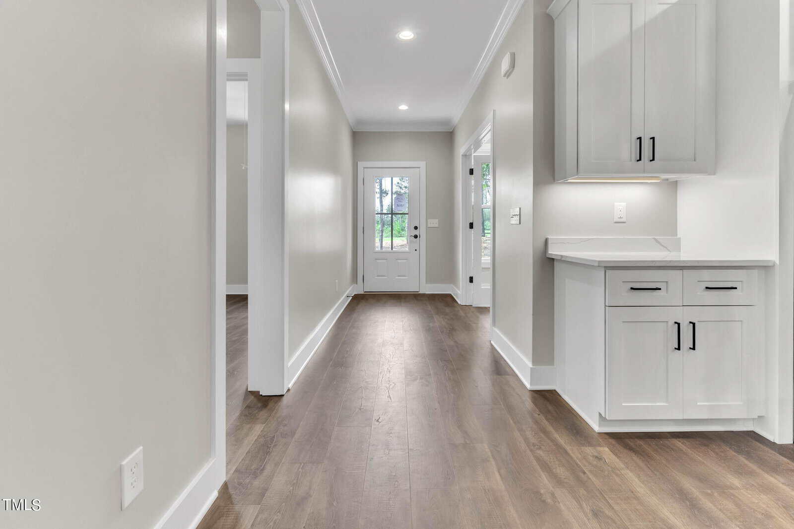 A bright hallway with light gray walls and wooden flooring leads to a white door with glass panels at the end. On the left, there are doorways to other rooms, while on the right, there are white cabinets with black handles. Recessed lighting illuminates the space.