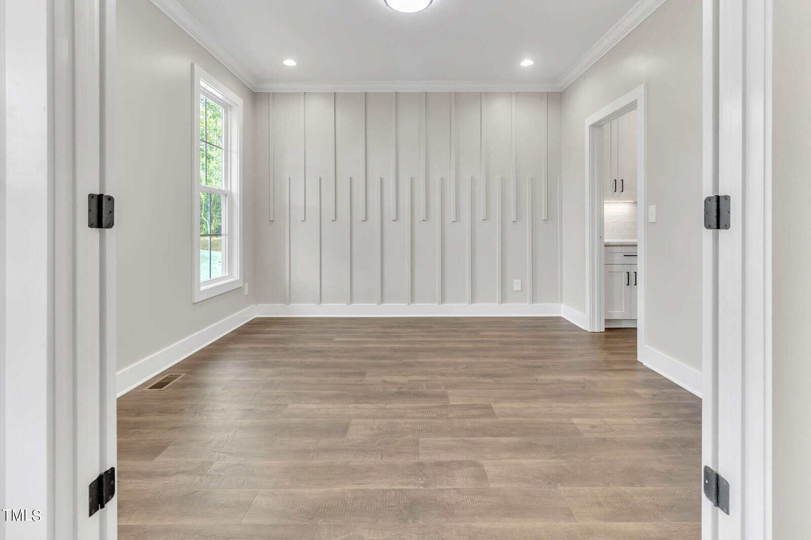 A modern, empty room with light gray walls and wooden flooring is shown. The room features a large window on the left, recessed lighting, and paneled wall decorations. A doorway leads to a kitchen area with white cabinetry.