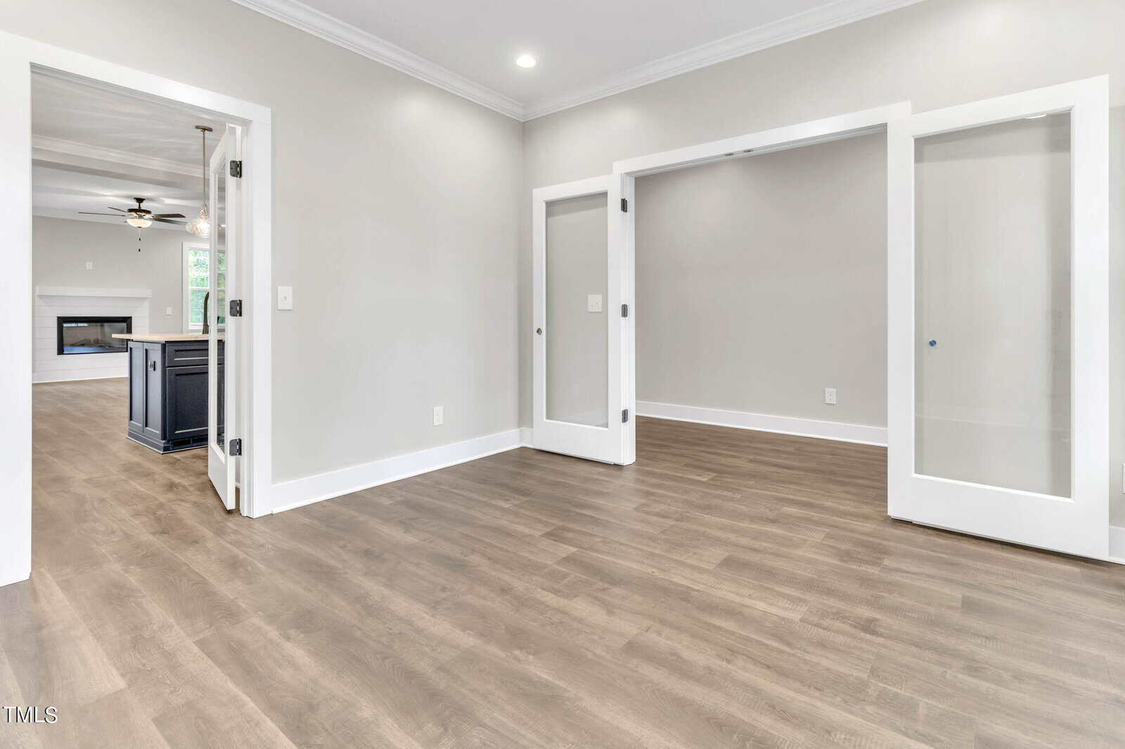 A bright, empty room with light gray walls and wood flooring. Two large sliding glass doors with white frames are partially open, revealing another empty room. In the background, a kitchen and living area with a ceiling fan and fireplace are visible.