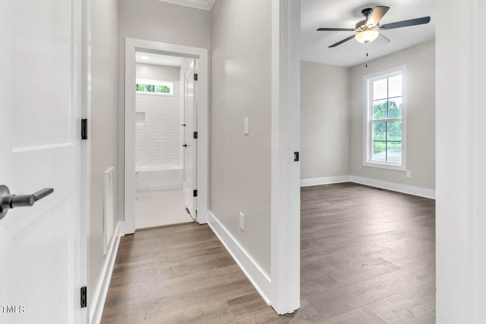 A hallway in a modern home features light gray walls and dark hardwood flooring. To the left is a closed white door, and straight ahead is a bathroom with white tile. To the right is a bedroom with large windows, dark hardwood floors, and a ceiling fan.