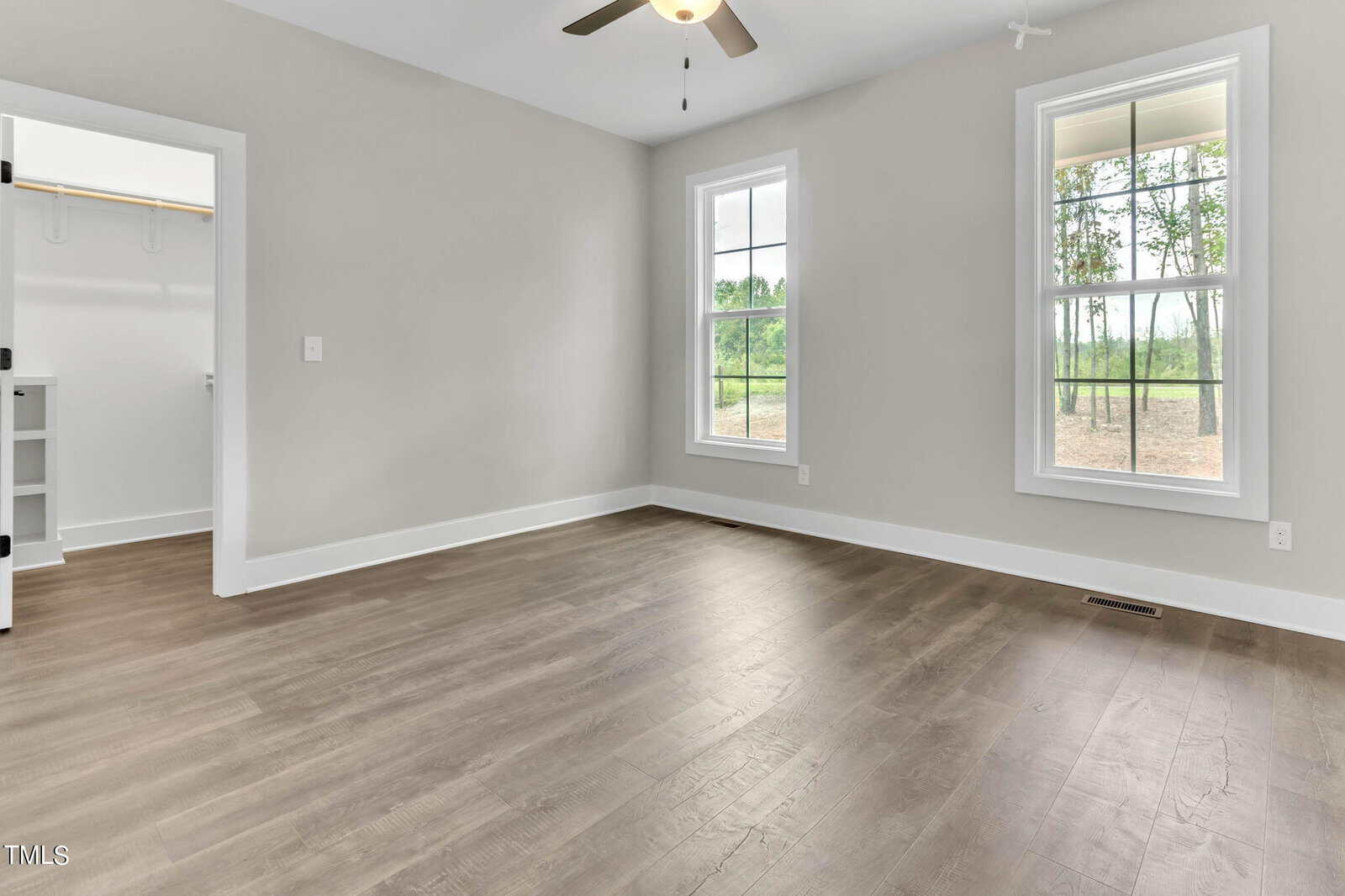 A bright and empty room with light gray walls, large windows, a ceiling fan with a light, and light-colored hardwood floors. There is a small open door leading to a closet with built-in shelves and hanging rods. Trees can be seen outside the windows.