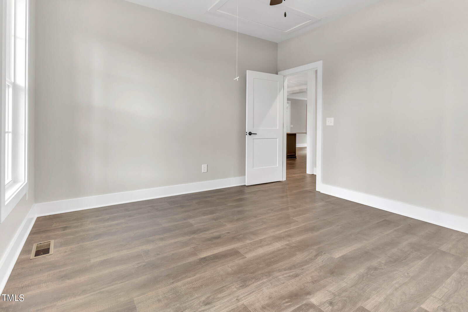 A vacant room with light gray walls and wood flooring. The room features a window on the left, a white framed door with a glass panel on the right, and white trim along the baseboards. The door opens into another area with hardwood floors visible.