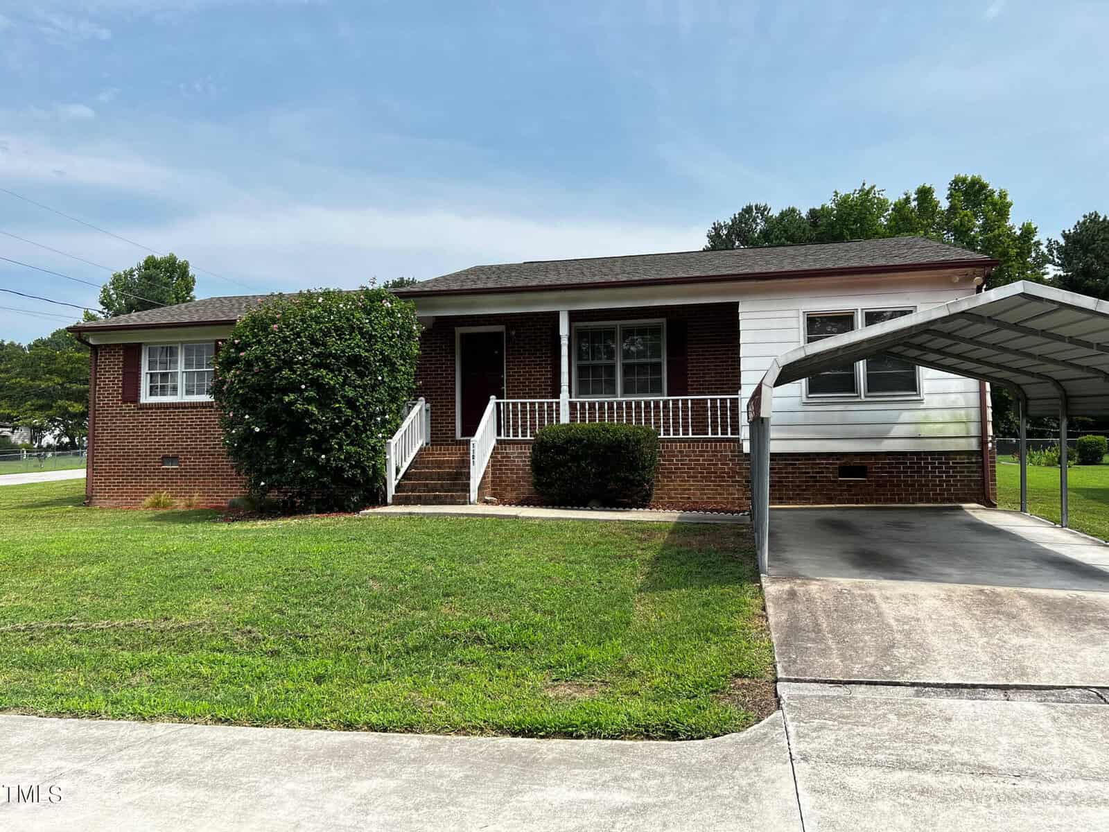 A small brick house with white siding, a front porch, and steps leading to the entrance sits nicely at 1101 Water Street in Henderson. It has a manicured lawn, a bush to the left of the steps, and a carport on the right. The driveway is in the foreground, and the sky is clear with a few clouds.